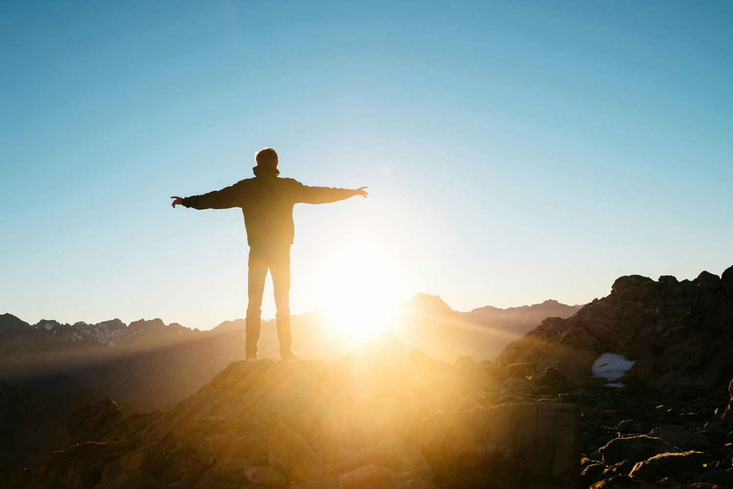 Person standing on a mountain peak with arms outstretched, silhouetted against sunrise.