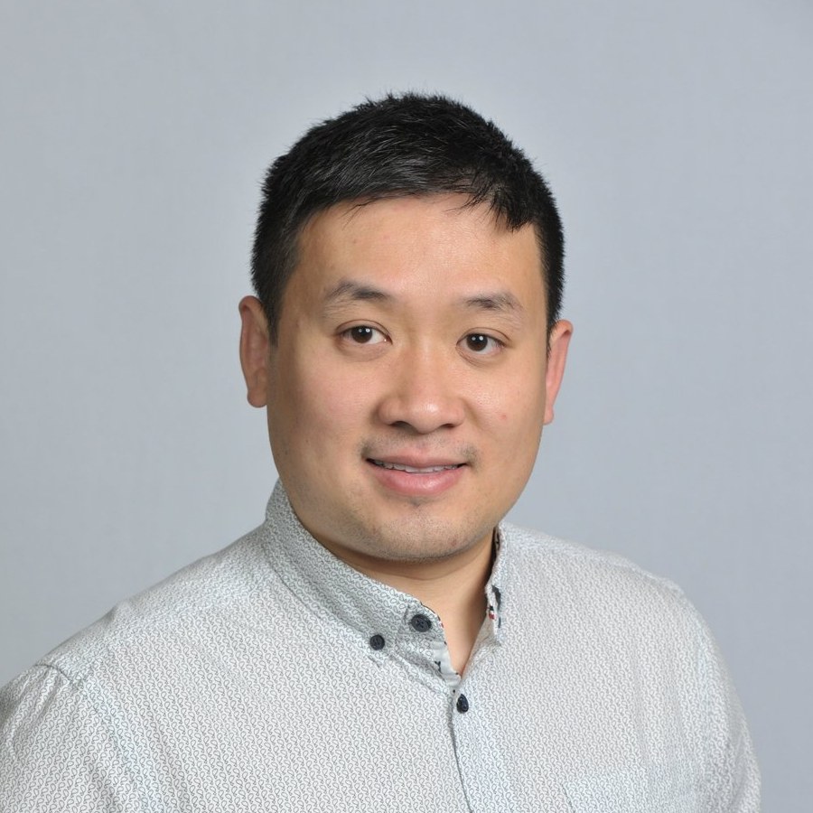 A young man with short black hair and light skin, smiling, wearing a light-colored, patterned button-up shirt, standing against a plain light gray background.